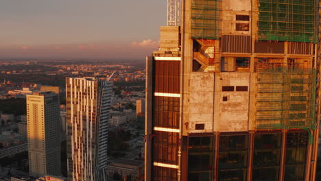 Fly-around-top-of-skyscraper-construction-site-high-above-city.-Concrete-walls-and-scaffolding-lit-by-setting-sun.-Warsaw,-Poland