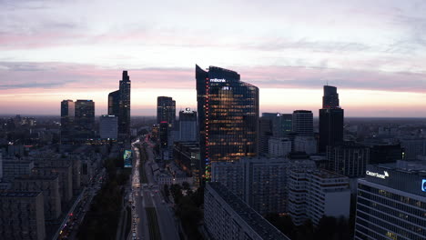 Modern-downtown-high-rise-buildings-against-twilight-sky.-Descending-shot-of-wide-busy-street-between-skyscrapers.-Warsaw,-Poland