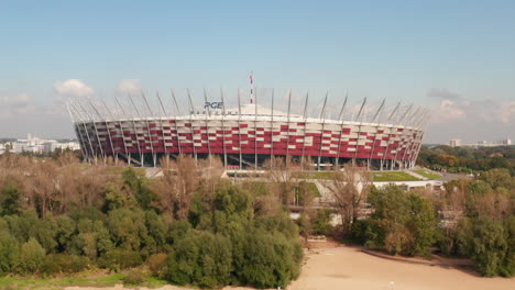 Fly-around-Polish-National-stadium,-modern-multifunction-arena.-Low-angle-view-above-trees-in-park.-Warsaw,-Poland