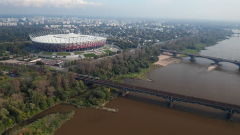 slide-and-pan-footage-of-modern-National-stadium-on-Vistula-riverbank.-Train-riding-on-bridge-over-water.-Warsaw,-Poland