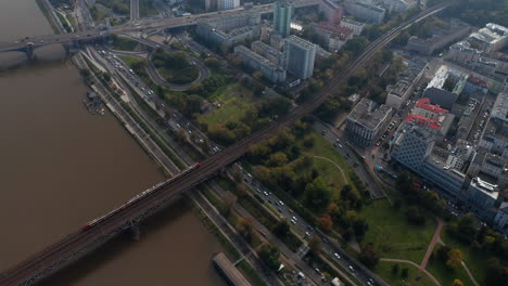 High-angle-view-of-traffic-in-city.-Tracking-of-passenger-train-running-on-track-across-busy-waterfront-road-and-Vistula-river.-Warsaw,-Poland