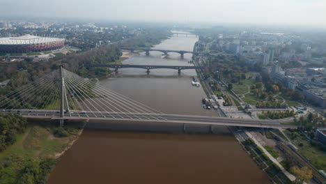 Forwards-fly-above-river.-Tilt-up-reveal-row-of-various-bridges-connecting-bank-in-city.-Modern-National-stadium-on-Vistula-riverbank.-Warsaw,-Poland
