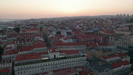 Aerial-view-of-colorful-rooftops-of-traditional-old-houses-in-Lisbon-city-center-with-reveal-of-Ponte-25-de-Abril-red-bridge-in-the-background