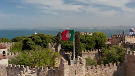 Close-up-of-national-and-municipal-flags-on-poles-over-medieval-stone-Saint-George-Castle.-Drone-camera-flying-backward-away-from-landmark.-Lisbon,-capital-of-Portugal.