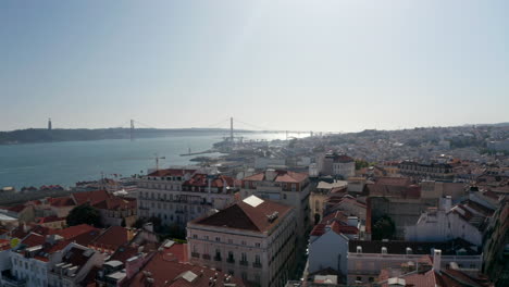 Panoramic-aerial-view-of-town-with-long-cable-stayed-bridge-spanning-Tagus-river.-Drone-camera-descending-between-buildings-and-gradually-hiding-view.-Lisbon,-capital-of-Portugal.