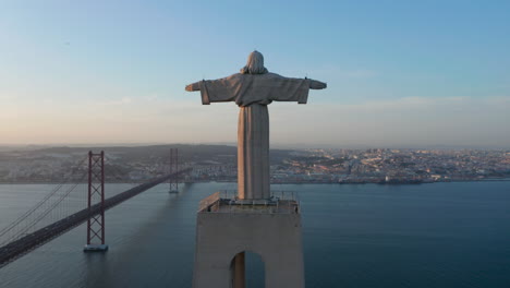 Drone-camera-ascending-along-pedestal-and-statue.-Evening-back-view-of-Christ-the-King-Sanctuary-in-Almada.-Long-cable-stayed-bridge-over-Tagus-river-in-background.-Lisbon,-capital-of-Portugal.