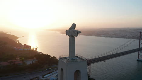 Stunning-aerial-orbit-of-Sanctuary-of-Christ-the-King-statue-on-the-hill-in-evening-light-with-reveal-of-Ponte-25-de-Abril-red-bridge-and-Lisbon-city-center-across-the-sea
