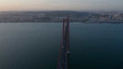 Evening-aerial-view-of-25th-of-April-Bridge-connecting-Lisbon-and-Almada.-Long-cable-stayed-highway-bridge-over-Tegus-river.-Drone-rotating-around.-Lisbon,-capital-of-Portugal.