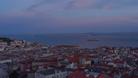 Aerial-view-of-evening-city-rooftops.-Drone-flying-over-red-roofs-after-sunset.-Large-water-surface-of-Tegus-river-in-background.-Lisbon,-capital-of-Portugal.