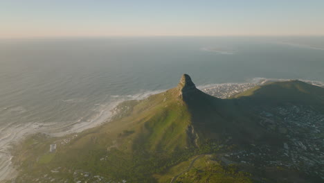 Aerial-footage-of-ocean-coast.-Pointed-mountain-towering-high-above-houses-in-urban-boroughs.-Cape-Town,-South-Africa