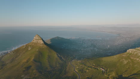 Aerial-panoramic-footage-of-metropolis-between-mountains-and-sea-bay.-Sun-shining-on-rock-top-of-Lions-Head-mountain-above-city.-Cape-Town,-South-Africa