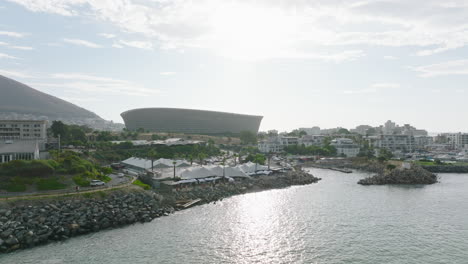 Diapositiva-Y-Toma-Panorámica-Del-Moderno-Estadio-De-Fútbol-De-Punto-Verde,-Volar-A-Lo-Largo-De-La-Costa-Del-Mar-Con-Restaurante.-Ciudad-Del-Cabo,-Sudáfrica