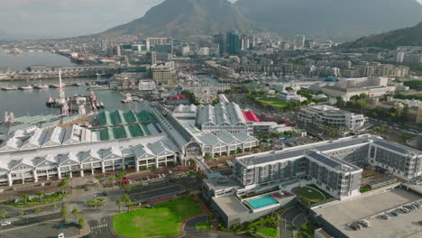 Aerial-descending-footage-of-shopping-and-entertainment-centre-Victoria-Wharf-on-waterfront.-Cityscape-and-high-mountains-in-distance.-Cape-Town,-South-Africa
