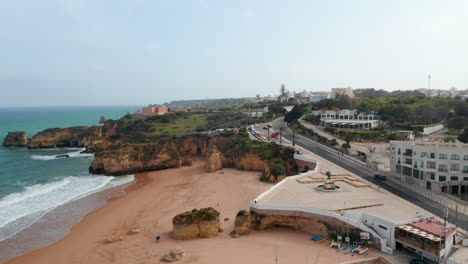 Aerial-drone-flying-forward-above-paradisiac-shoreline-beach-coast-in-Lagos,-Algarve,-cyclist-passing-by-on-pier,-Portugal,-day