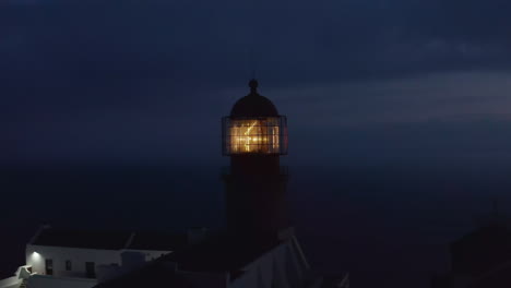 Leuchtendes-Leuchtturm-scheinwerferlicht-In-Der-Dämmerung,-Drohne-Kreist-Herum-Mit-Hintergrundabendmeer,-Lagos,-Portugal