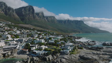 Aerial-footage-of-properties-at-sea-coast.-Waves-washing-coastal-rocks.-Majestic-mountain-ridge-shrouded-in-clouds-in-background.-Cape-Town,-South-Africa