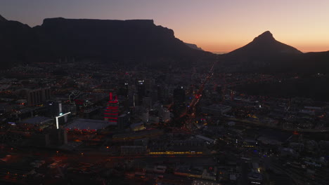 City-in-evening.-Aerial-footage-of-illuminated-buildings-and-streets-with-traffic-after-sunset.-Silhouette-of-Table-Mountain-in-background.-Cape-Town,-South-Africa