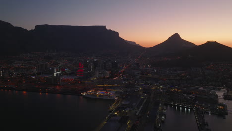 Aerial-shot-of-city-at-twilight.-Large-cruise-boat-mooring-at-waterfront.-Mountains-against-colourful-sky-in-background.-Cape-Town,-South-Africa