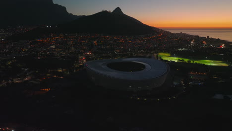 Imágenes-Aéreas-De-Diapositivas-Y-Panorámicas-De-La-Ciudad-Con-Un-Estadio-Deportivo-Moderno.-Vista-Panorámica-De-La-Ciudad-Junto-Al-Mar-Y-Las-Montañas-Contra-El-Cielo-Crepuscular.-Ciudad-Del-Cabo,-Sudáfrica