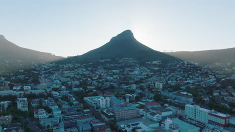 Tall-spiky-peak-casting-shadow-on-residential-borough-in-city.-Aerial-ascending-footage-revealing-bright-sun-behind-Lions-Head-mountain.-Cape-Town,-South-Africa
