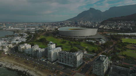 Aerial-ascending-footage-of-modern-football-arena-in-city.-Green-Point-Stadium-near-sea-coast.-Mountains-in-background.-Cape-Town,-South-Africa