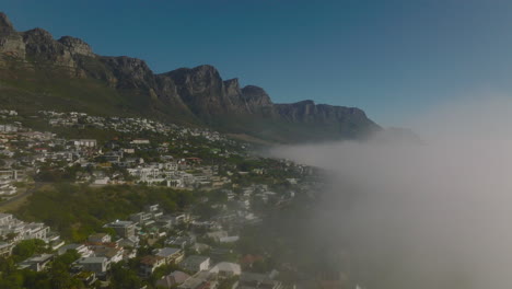 Forwards-fly-along-fog-rising-from-sea-along-coast.-Residential-houses-in-slope-under-rocky-escarpment.-Cape-Town,-South-Africa