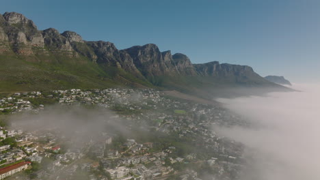 Descending-shot-of-rock-escarpment-above-tourist-destination.-Fog-rising-from-sea.-Cape-Town,-South-Africa
