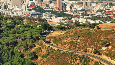 Vista-De-ángulo-Alto-De-Los-Vehículos-Que-Conducen-Por-Carretera-En-La-Naturaleza.-Incline-Hacia-Arriba-La-Revelación-De-Edificios-En-Una-Gran-Ciudad.-Ciudad-Del-Cabo,-Sudáfrica