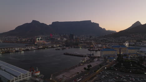 Backwards-reveal-of-sea-coast-in-town.-Marina-and-buildings-on-waterfront,-silhouette-of-Table-Mountain-against-colourful-twilight-sky.-Cape-Town,-South-Africa