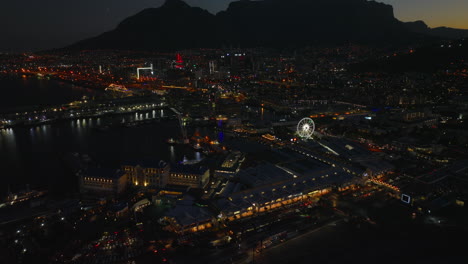 High-angle-view-of-waterfront-with-marina-at-night.-Tilt-up-reveal-of-city-and-silhouette-of-mountain-ridge-in-background.-Cape-Town,-South-Africa