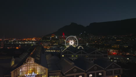 Forwards-ascending-fly-above-buildings-in-city-at-night.-Revealing-Ferris-wheel-and-silhouette-of-mountain-ride-in-background.-Cape-Town,-South-Africa