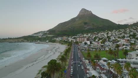 Forwards-fly-above-coastal-road-leading-between-buildings-and-sand-beach-at-twilight.-Lions-Head-mountain-in-background.-Cape-Town,-South-Africa