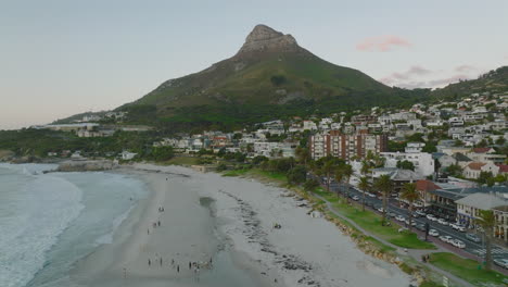Aerial-shot-of-people-enjoying-evening-on-sand-beach.-Buildings-in-urban-borough-on-seaside-at-twilight.-Cape-Town,-South-Africa