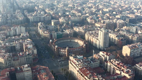 Forwards-fly-above-city.-Famous-historical-landmark,-La-Monumental-arena-and-surrounding-buildings-lit-by-bright-low-sun.-Barcelona,-Spain