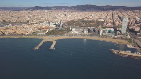 Aerial-panoramic-footage-of-sea-coast-and-metropolis.-Various-buildings-in-large-city-lit-by-bright-sun-and-hills-in-background.-Barcelona,-Spain