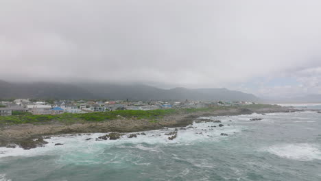 Slide-and-pan-shot-of-waves-crashing-on-rocks-at-sea-coast.-Family-houses-in-town-in-background.