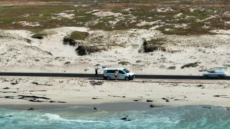 Slide-and-pan-footage-of-passenger-van-standing-next-to-road-on-sea-coast.-Waves-washing-sand.-South-Africa