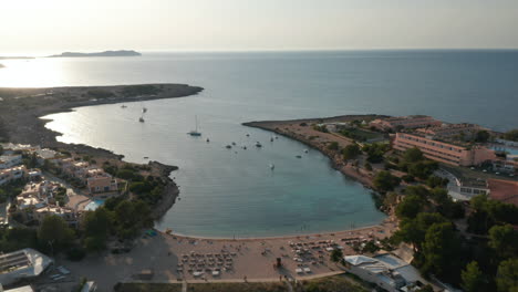 Luftpanorama-Zeitlupenansicht-Einer-Menge-Nicht-Erkennbarer-Menschen,-Die-Sich-Am-Strand-Entspannen-Und-Genießen,-Umgeben-Von-Resort-Und-Meer-Mit-Bergen,-Während-Des-Abends-Auf-Ibiza-In-Spanien