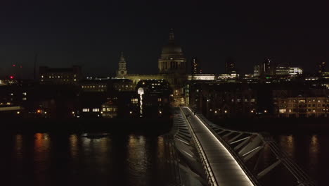 People-walking-across-Millennium-Bridge-at-night.-View-of-modestly-illuminated-Saint-Pauls-Cathedral.--London,-UK