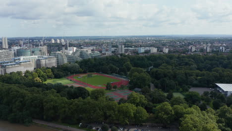 Forward-fly-towards-Battersea-Park.-Aerial-view-of-sport-areas-and-urban-neighbourhood-behind.-London,-UK