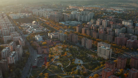 Aerial-shot-of-park-surrounded-by-multistorey-apartment-houses-on-housing-estate.-Colourful-trees-lit-by-setting-sun.