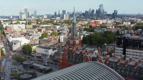 Orbit-shot-around-clock-tower-at-old-brick-building-of-St-Pancras-train-station.-Aerial-panoramic-view-of-city-with-tall-skyscrapers-in-distance.-London,-UK