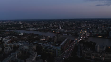Forward-fly-above-evening-city.-Aerial-view-at-illuminated-famous-Tower-Bridge-over-River-Thames.-Dark-scene-after-sunset.-London,-UK
