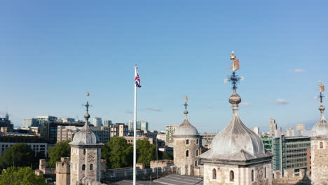 Panorama-curve-shot-of-top-of-Tower-of-London.-Close-up-of-four-towers-and-Union-Jack-on-pole-above-roof-of-stone-keep.-Modern-business-centre-in-background.-London,-UK