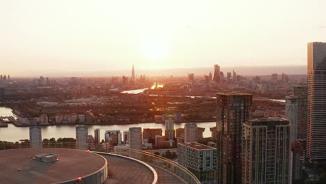 Aerial-panoramic-view-of-city-against-setting-sun,-downtown-skyscrapers-in-distance.-Descending-footage-reveals-top-of-Arena-Tower-cylindrical-apartment-building.-London,-UK