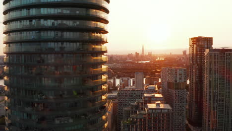 Vuele-Alrededor-De-La-Torre-De-Arena-De-Un-Edificio-Residencial-Cilíndrico-Alto-Y-Moderno-En-El-Distrito-De-Canary-Wharf.-Distrito-De-La-Ciudad-Futurista-En-El-Atardecer.-Londres,-Reino-Unido