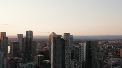 Fly-around-tall-skyscrapers-towering-high-above-other-buildings-in-city.-Parallax-effect.-Evening-aerial-footage-of-Canary-Wharf.-London,-UK
