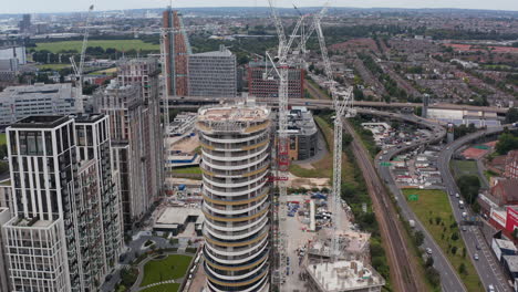 Tilt-and-pan-shot-of-modern-design-tall-building-construction-site.-Tall-tower-cranes-transporting-material.-New-residential-complex-at-White-city-borough.-London,-UK