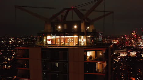 Orbit-shot-around-illuminated-top-of-modern-tall-building.-Night-cityscape-moving-in-background.-London,-UK