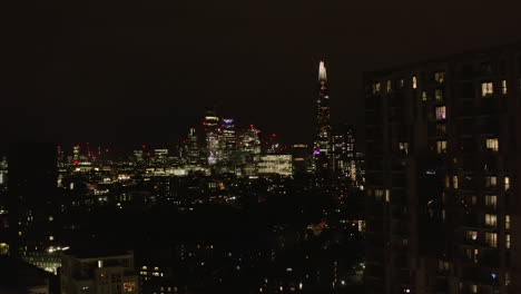 Forwards-fly-above-urban-neighbourhood-at-night.-Revealing-view-of-glowing-downtown-skyscrapers.-London,-UK